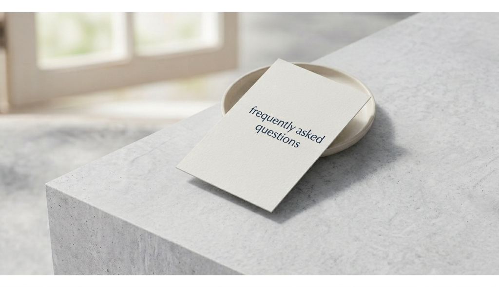 A minimalist cream-colored card resting on a small ceramic dish over a light gray textured concrete surface. The card cleanly displays the text 'frequently asked questions' in a modern navy blue lowercase font under soft natural lighting